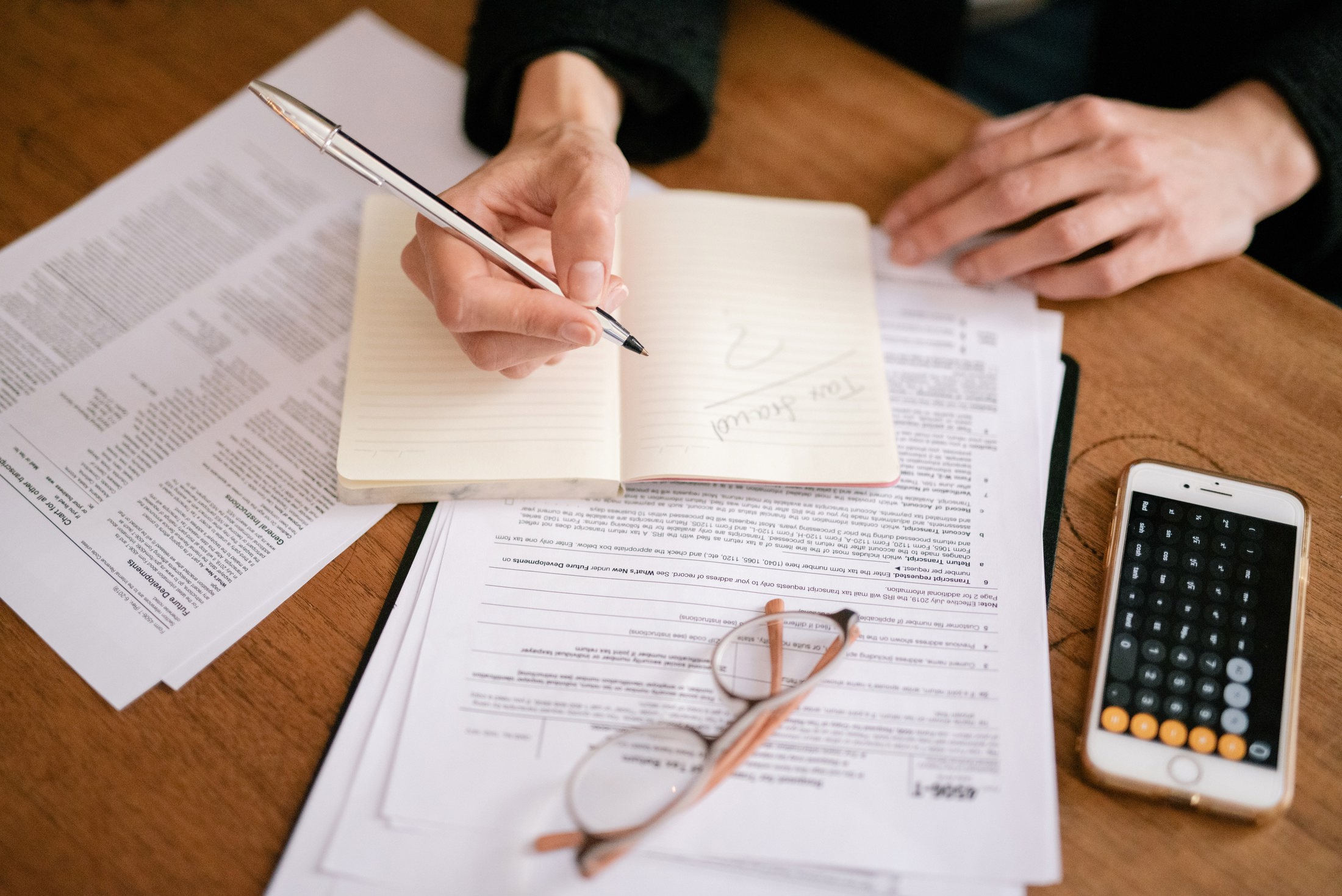 A woman's hands writing in a notepad on top of papers, beside a smartphone calculator and glasses